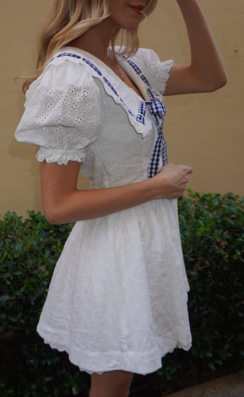 Woman wearing a white dress with a blue trim against a beige wall and green bushes.